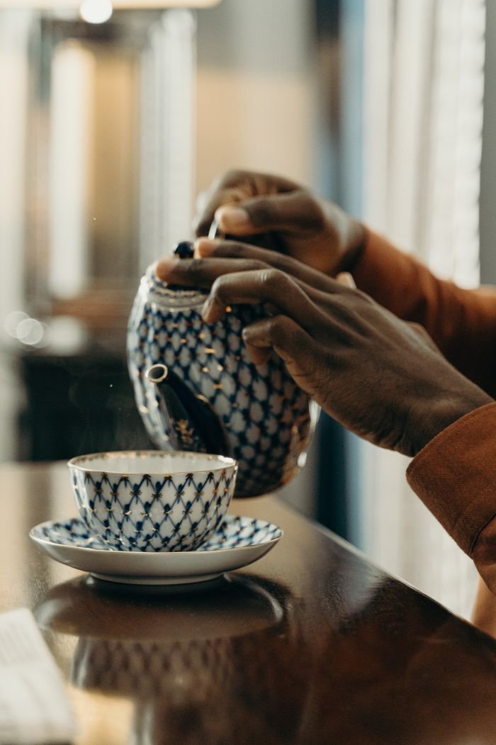 A person pours tea from a decorative teapot into a porcelain cup, captured in a cozy indoor setting.
